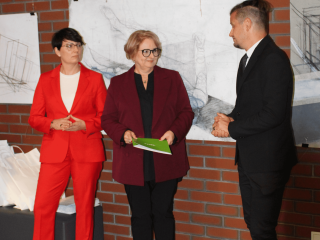 Dwie kobiety i mężczyzna elegancko ubrani, stojący na tle ściany z czerwonych cegieł.