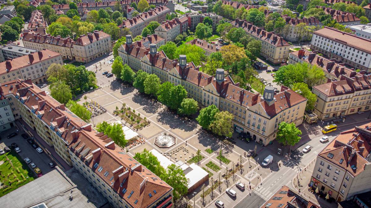 Widok na plac otoczony budynkami z dachem z czerwonej cegły,  wokół liczne drzewa, a w centralnym punkcie fontanna.