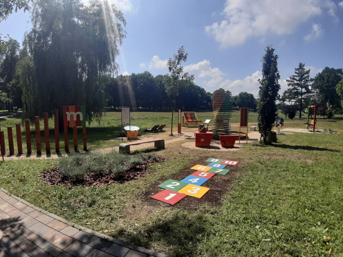 plac zabaw z namalowanymi kolorowymi polami z cyframi, wokół zieleń, drzewa, u góry błękitne niebo