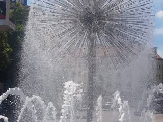 Tryskająca woda z fontanny, w oddali brukowany plac i budynki mieszkalne